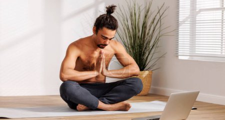Téléchargez les photos : Jeune homme afro-américain joyeux avec torse nu assis dans la pose de lotus devant un ordinateur portable et remercie professeur dans le salon à la maison, ayant cours de yoga en ligne. Regarder des tutoriels de yoga en ligne - en image libre de droit