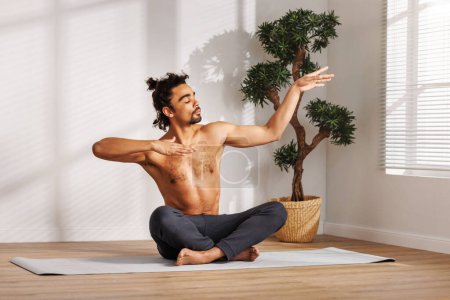 Photo for Shirtless black man with closed eyes gesticulating while sitting in Padmasana pose near potted tree and meditating during yoga session in morning at home - Royalty Free Image