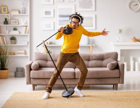 Photo for Full body African American male in casual clothes listening to music in wireless headphones and singing into lamp while standing near sofa in light living room at home - Royalty Free Image