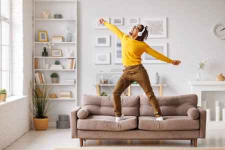 Téléchargez les photos : Homme noir complet en vêtements décontractés et écouteurs sans fil levant les bras, sautant et dansant sur le canapé tout en s'amusant dans le salon le week-end à la maison - en image libre de droit