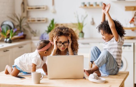 Photo for Young irritated stressed African-American mother covering ears and looking at laptop screen while working remotely from home with two small kids, being distracted from work by noisy naughty children - Royalty Free Image