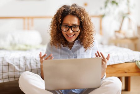 Photo for Expressive emotional african american teenage girl sitting near bed at home smiling and gesticulating during a video call to a friend via laptop  with excited expression - Royalty Free Image