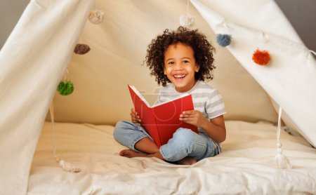 Photo for Portrait of cute little african american kid curly boy with book smiling at camera while   reading book  in play tent at home, happy child playing alone in children room - Royalty Free Image