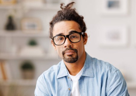 Foto de Retrato de un serio empresario afroamericano en gafas y con el pelo largo mirando a la cámara en la oficina en casa sobre un fondo borroso - Imagen libre de derechos