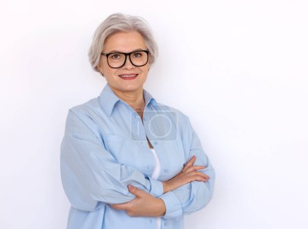 Téléchargez les photos : Enthousiaste femme entrepreneur mature avec les cheveux gris et dans des lunettes debout avec les bras croisés sur fond blanc et regardant la caméra - en image libre de droit