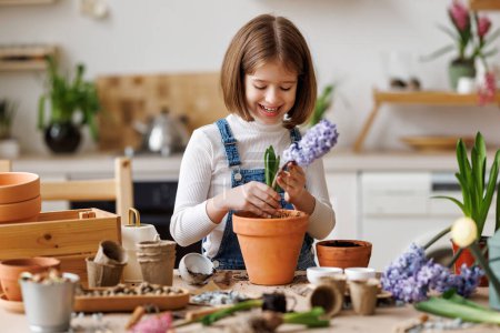 Foto de Niño alegre en ropa casual con pelo corto sonriendo y plantando flor de jacinto violeta en maceta mientras hace jardinería en casa en la mesa en la cocina - Imagen libre de derechos