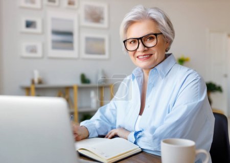 Foto de Mujer empresaria sonriente de mediana edad que trabaja en la computadora portátil, lee documentos, toma notas y analiza informes mientras trabaja en proyectos en el lugar de trabajo doméstico - Imagen libre de derechos