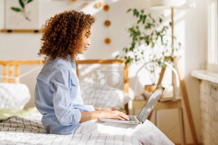 Foto de Sonriente joven mujer afroamericana con peinado afro sentado en la cama mensajería con un amigo en el ordenador portátil o ver videos divertidos mientras pasa el tiempo libre y relajarse en el acogedor dormitorio de luz en casa - Imagen libre de derechos