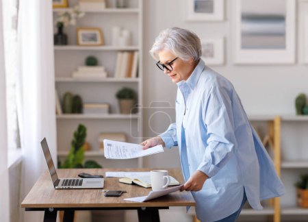Photo for Focused middle aged female entrepreneur working at laptop, reading documents,  take notes and analyzing reports while working on project in domestic workplace - Royalty Free Image