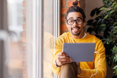 Téléchargez les photos : Homme noir en vêtements décontractés et lunettes parler tout en faisant appel vidéo via tablette près de la fenêtre dans la journée à la maison - en image libre de droit