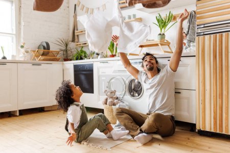Photo for Happy african american dad and son smiling and tossing towels in air while sitting on floor near washing machine during household routine in kitchen at home - Royalty Free Image