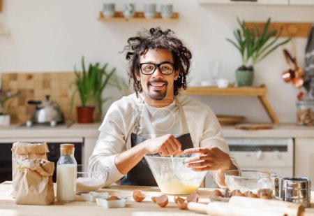 Foto de Sonriente macho afroamericano en delantal añadiendo ingredientes en un tazón mientras hace masa para cocinar panadería en la cocina en casa - Imagen libre de derechos