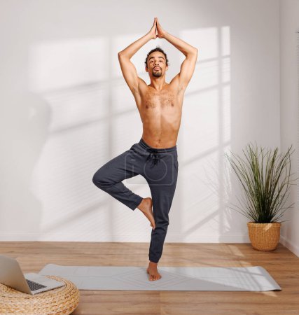 Photo for Calm African American man with naked torso standing  in Tree Vrksasana pose near potted tree and meditating with closed eyes in morning in sunlit room at home - Royalty Free Image