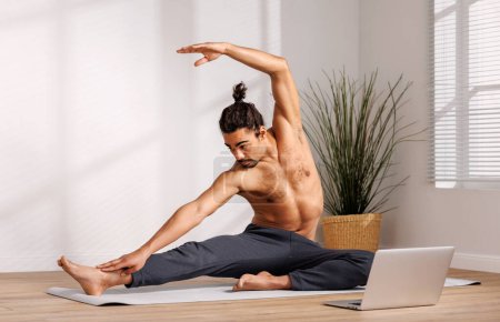Foto de Hombre afroamericano sin camisa con el moño del pelo levantando el brazo y doblando a un lado mientras que está sentado en la estera cerca del ordenador portátil y haciendo yoga en la mañana en casa - Imagen libre de derechos