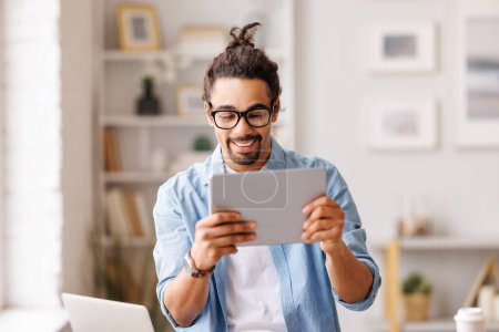 Photo for Cheerful African American male hipster in glasses browsing tablet and working remotely on project in home workspace - Royalty Free Image