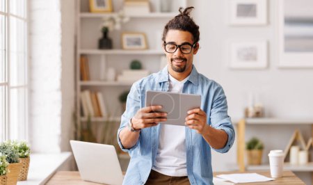 Téléchargez les photos : Joyeux hipster homme afro-américain dans des lunettes de navigation tablette et de travailler à distance sur le projet dans l'espace de travail à domicile - en image libre de droit