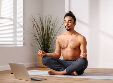 Photo for Calm African American man with naked torso sitting in Lotus pose  in front of laptop   in living room at home, having yoga class online and meditating with closed eyes in morning in sunlit room - Royalty Free Image