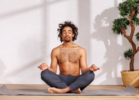 Foto de Hombre afroamericano tranquilo con el torso desnudo sentado en la pose del loto cerca del árbol en maceta y meditando con los ojos cerrados en la mañana en la habitación soleada en casa - Imagen libre de derechos