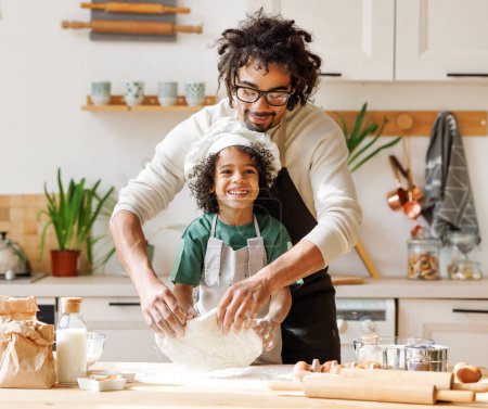 Téléchargez les photos : Homme afro-américain joyeux et garçon dans des tabliers faire de la pâtisserie à la table en bois dans la cuisine tout en cuisinant la boulangerie ensemble - en image libre de droit