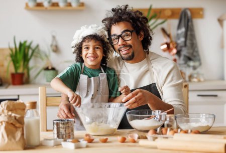 Foto de Contenido cariñoso padre afroamericano y lindo niño añadiendo ingredientes en el tazón y preparando la masa en la mesa en casa - Imagen libre de derechos