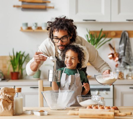 Téléchargez les photos : Contenu aimant père afro-américain et mignon garçon ajoutant des ingrédients dans le bol et la préparation de la pâte à table à la maison - en image libre de droit