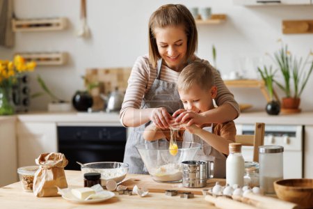 Content loving mother and cute boy adding ingredients in bowl and preparing dough at table at home