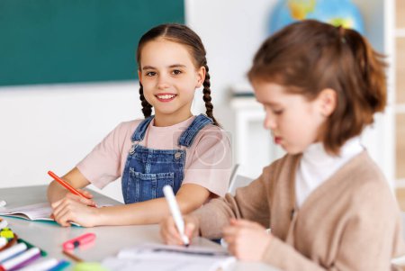 Photo for Gleeful school girl in casual clothes smiling and looking at camera while sitting at desk near classmate and drawing in notepad during lesson in daytime at school - Royalty Free Image