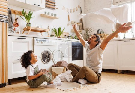 Téléchargez les photos : Heureux père afro-américain et fils souriant et jetant des serviettes dans l'air tout en étant assis sur le sol près de la machine à laver pendant la routine domestique dans la cuisine à la maison - en image libre de droit