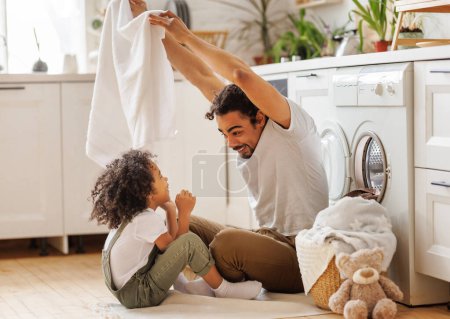 Foto de Optimista negro familia padre y feliz hijo en casual ropa sonriendo y cubriéndose con toalla mientras están sentados en el suelo y haciendo lavandería en la cocina en casa - Imagen libre de derechos