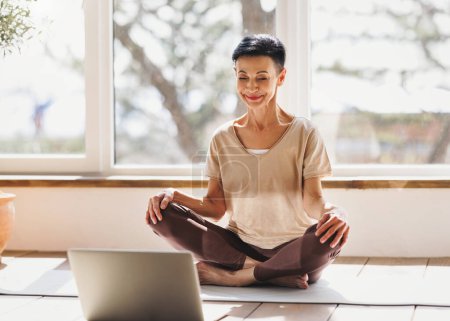 Téléchargez les photos : Femme d'âge moyen pieds nus en vêtements de sport fermer les yeux et méditer avec le sourire dans Lotus pose contre la fenêtre lors d'une séance de yoga en ligne le matin à la maison - en image libre de droit