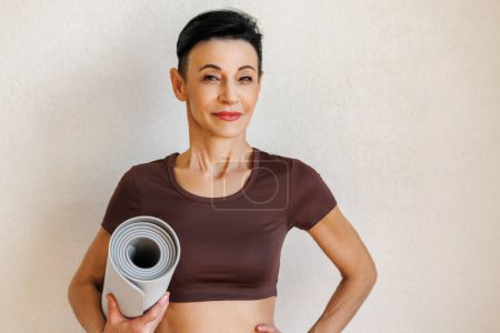 Photo for Glad mature female with rolled up mat holding hand on waist and looking at camera with smile while standing against beige wall before fitness training - Royalty Free Image