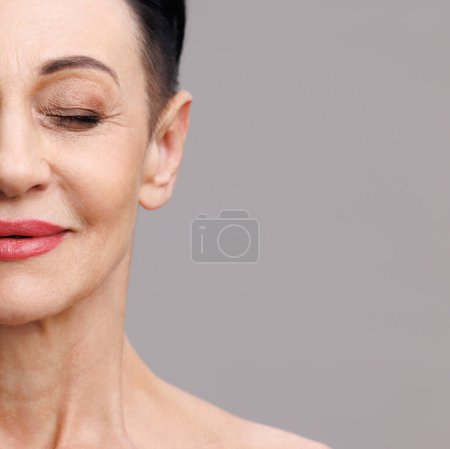 Téléchargez les photos : La moitié du visage d'une femme mature avec un maquillage élégant fermant les yeux et souriant pendant la routine de beauté sur fond gris - en image libre de droit
