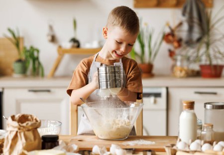 Foto de Sonriente niño lindo en delantal la adición de ingredientes en el tazón, mientras que la fabricación de masa para cocinar panadería en la cocina en casa - Imagen libre de derechos
