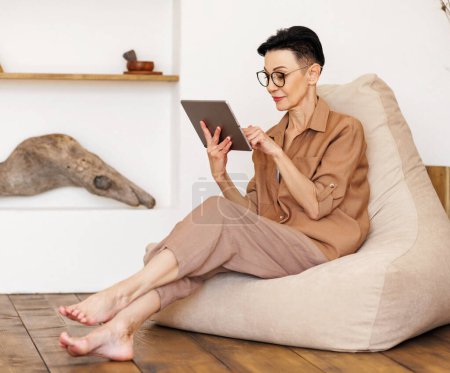 Photo for Barefoot mature female in casual clothes and glasses browsing social media on tablet while resting on bean bag near shelves with decorations in light room at home - Royalty Free Image