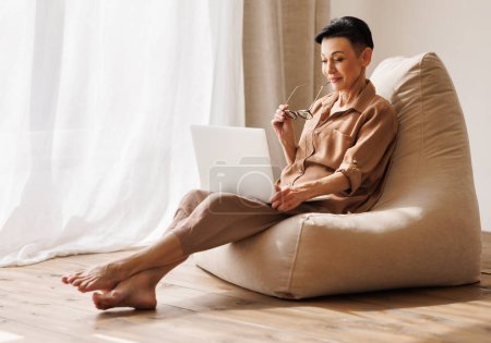 Photo for Barefoot mature female in casual clothes and glasses browsing social media on laptop while resting on bean bag near shelves with decorations in light room at home - Royalty Free Image