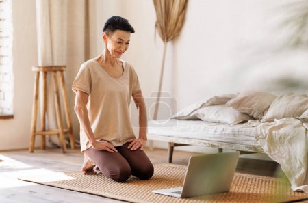 Foto de Mujer madura positiva con cabello corto y oscuro arrodillado en el suelo cerca de la cama sin hacer y mirando la pantalla del portátil al inicio de la sesión de yoga en línea por la mañana en casa - Imagen libre de derechos