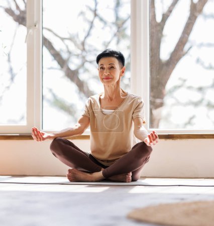 Photo for Ground level of relaxed mature woman with short dark hair sitting in Padmasana pose near window and meditating early in morning at home - Royalty Free Image
