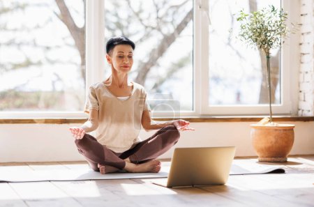 Foto de Mujer descalza de mediana edad en ropa deportiva cerrando los ojos y meditando en Lotus posan contra la ventana durante la sesión de yoga en línea por la mañana en casa - Imagen libre de derechos