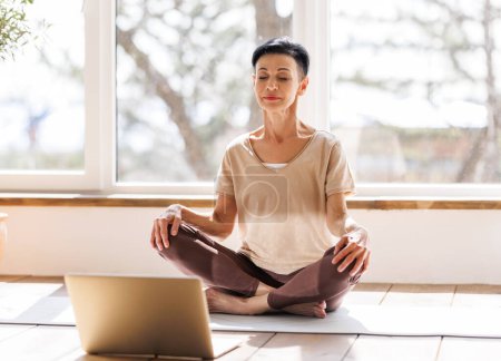 Téléchargez les photos : Femme d'âge moyen pieds nus en vêtements de sport fermer les yeux et méditer dans Lotus pose contre la fenêtre lors d'une séance de yoga en ligne le matin à la maison - en image libre de droit