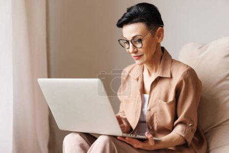 Foto de Mujer madura en ropa casual inteligente y gafas sentadas en bolsa de frijoles y portátil de navegación durante el día en casa - Imagen libre de derechos