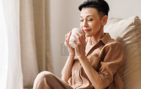 Photo for Middle aged female with short dark hair enjoying smell of fresh coffee with closed eyes while resting on bean bag in morning at home - Royalty Free Image