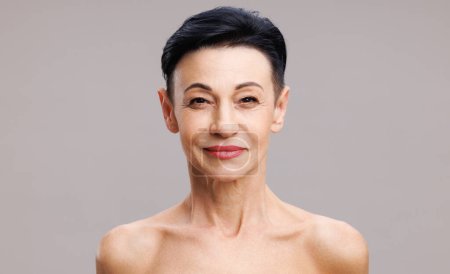 Foto de Cara de mujer madura con maquillaje elegante sonriendo durante la rutina de belleza contra fondo gris - Imagen libre de derechos