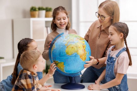Téléchargez les photos : Enseignant en lunettes et enfants positifs examinant globe tout en se tenant autour de la table dans la classe de géographie pendant la leçon à l'école - en image libre de droit
