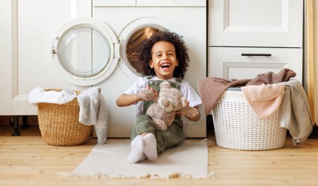 Photo for Happy laughing black boy with curly hair sitting on floor near  washing machine while doing laundry in kitchen at home - Royalty Free Image