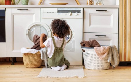 Téléchargez les photos : Vue arrière du garçon noir avec les cheveux bouclés agenouillés sur le sol et mettre des vêtements dans la machine à laver tout en faisant la lessive dans la cuisine à la maison - en image libre de droit