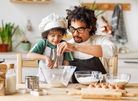 Foto de Contenido cariñoso padre afroamericano y lindo niño añadiendo ingredientes en el tazón y preparando la masa en la mesa en casa - Imagen libre de derechos