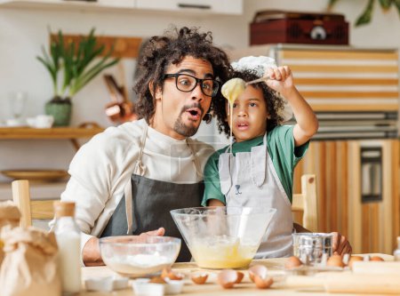 Foto de Sorprendido hombre y niño afroamericano en delantales preparando masa en un tazón mientras cocinan juntos en la mesa en la cocina - Imagen libre de derechos