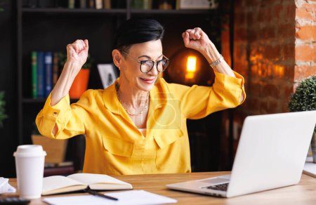 Foto de Mujer de negocios de mediana edad feliz con camisa amarilla de moda y gafas levantando los brazos y mirando a la pantalla del ordenador portátil con sonrisa después de terminar el proyecto mientras está sentado en el escritorio en la oficina en casa - Imagen libre de derechos