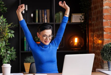 Téléchargez les photos : Heureuse femme d'affaires d'âge moyen en col roulé bleu tendance et lunettes levant les bras et regardant l'écran d'ordinateur portable avec le sourire après avoir terminé le projet tout en étant assis au bureau à la maison - en image libre de droit