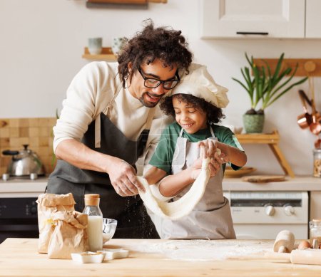 Foto de Alegre hombre y niño afroamericano en delantales haciendo pastelería en la mesa de madera en la cocina mientras cocinan panadería juntos - Imagen libre de derechos
